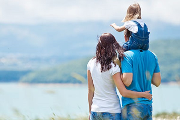 Mother and father stood in front of mountains and lake.  Father has a litle girl on his shoulders